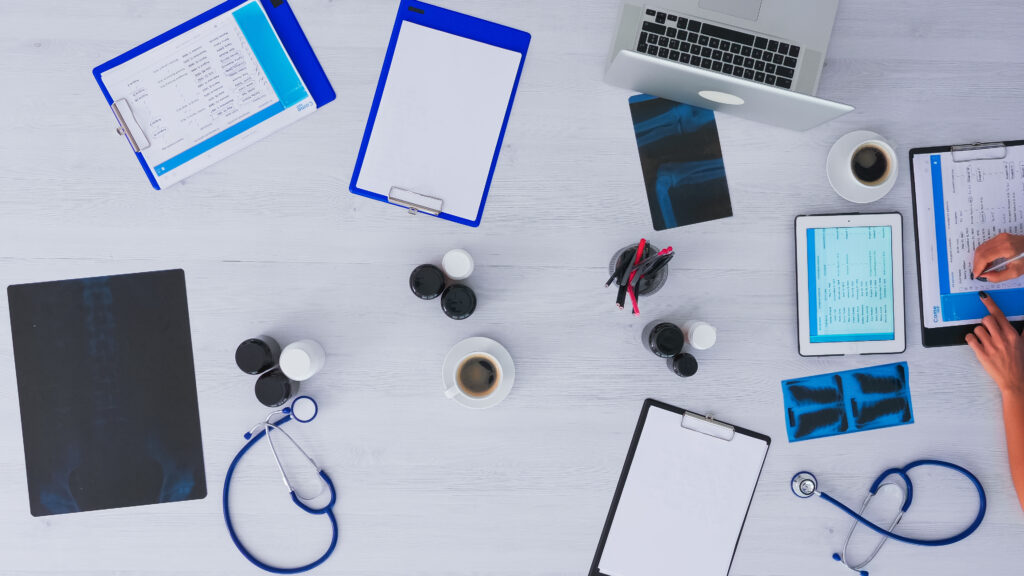 Medical workspace with EHR systems, patient records on clipboards, X-ray images, a laptop, a tablet, coffee, and a stethoscope, illustrating efficient healthcare management.