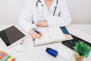 Medical professional in white coat writing notes in a notebook, surrounded by tablet, inhaler, water glass, and plant, symbolizing efficient dermatology billing and medical practice management.