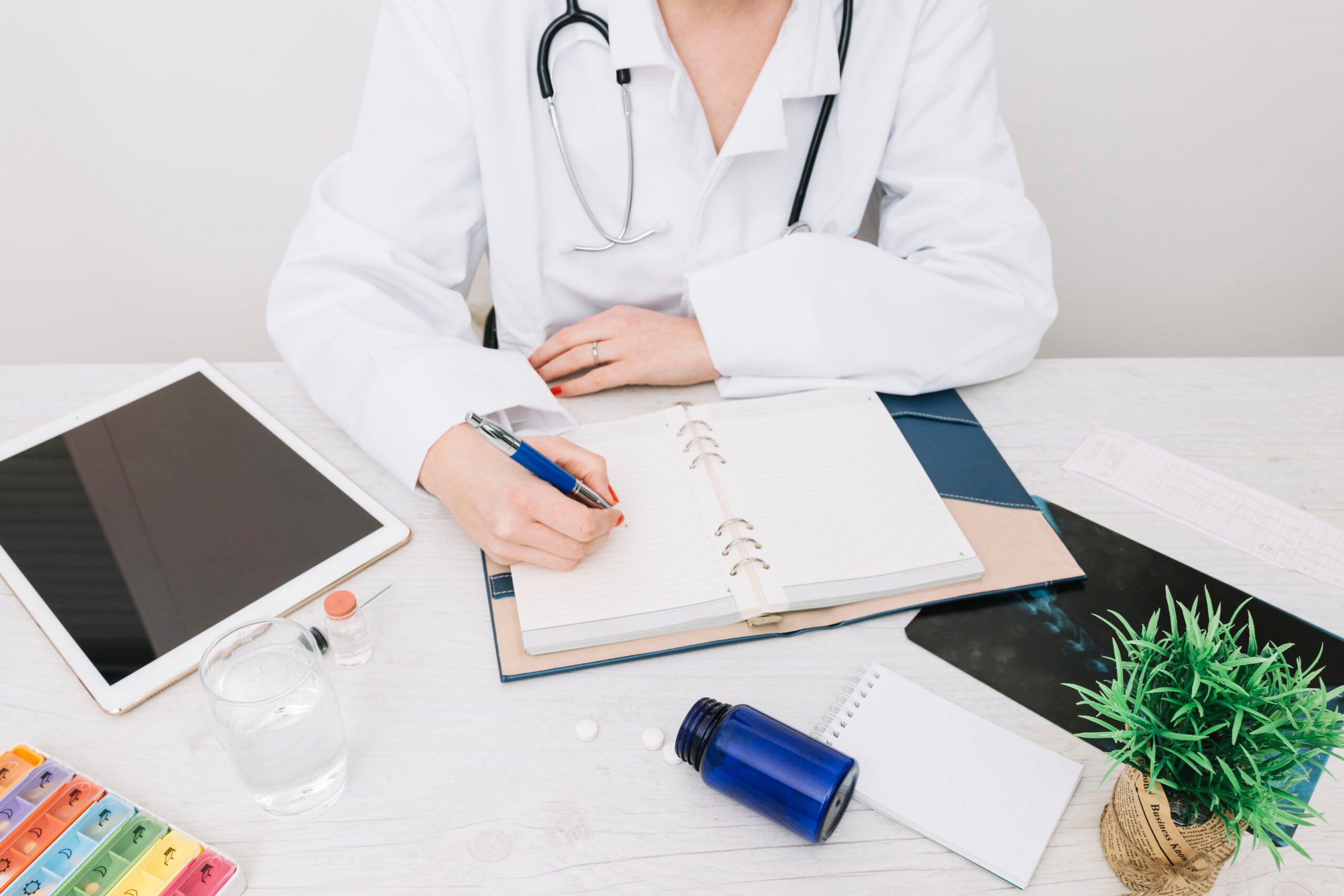 Doctor in a white coat writing notes in a notebook, with a stethoscope, tablet, glass of water, and medication on a desk, representing efficient dermatology billing practices.